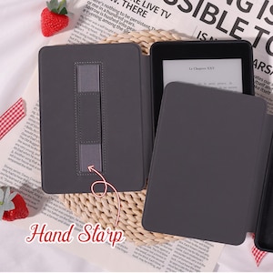 May include: A dark grey e-reader case with a hand strap is open, displaying the e-reader screen. The case rests on a patterned surface with text in the background. The text "Le Chapitre XXV" is visible on the screen.