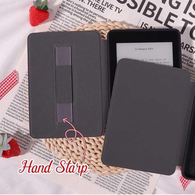 May include: A dark grey e-reader case with a hand strap is open, displaying the e-reader screen. The case rests on a patterned surface with text in the background. The text "Le Chapitre XXV" is visible on the screen.