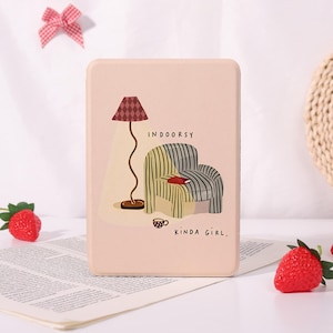 May include: A rectangular light pink e-reader case featuring a graphic illustration of a floor lamp, a striped armchair, a book, and a small cup. The text "INDOORSY KINDA GIRL." is printed in black lettering on the front of the protective cover.