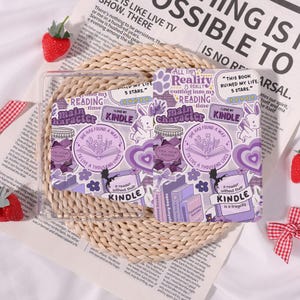May include: Clear phone case with purple stickers featuring book-themed designs, rabbits, hearts, and text like "I love my Kindle" and "Main Character." The case is on a woven mat with strawberries.