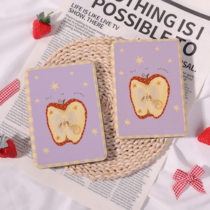 May include: Two rectangular coasters with a pale purple background and a whimsical apple design. The apple is cut in half, revealing the inside, and surrounded by yellow stars. The text "full of whimsy" is above the apple.