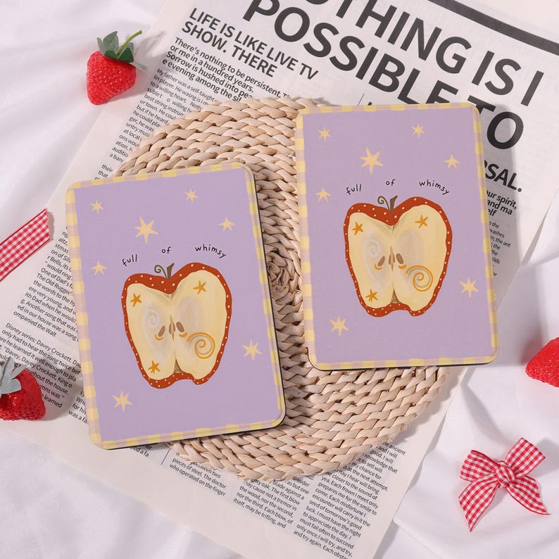 May include: Two rectangular coasters with a pale purple background and a whimsical apple design. The apple is cut in half, revealing the inside, and surrounded by yellow stars. The text "full of whimsy" is above the apple.