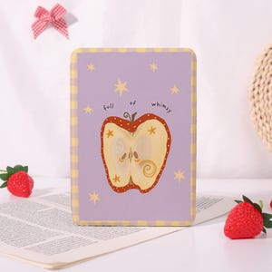 May include: A rectangular, lilac-coloured decorative sign featuring a whimsical apple illustration. The apple is halved, showing seeds and a swirl design. The text "full of whimsy" is above the apple. The sign has yellow stars and a yellow chequered border.