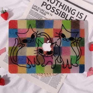 May include: A colorful laptop case featuring a grid pattern with cartoon cats. The case has a cutout for the Apple logo. The cats are outlined in black and colored with various hues. The background includes text that reads "NOTHING IS POSSIBLE."