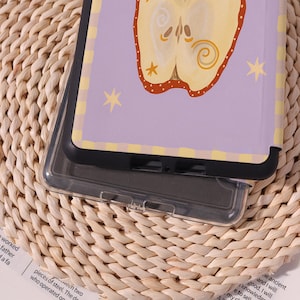 May include: A clear phone case with a purple and yellow design. The case features a stylized apple illustration with star accents. The case is resting on a woven surface.