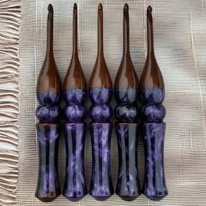 May include: Five handcrafted crochet hooks. The hooks have dark brown wooden shafts and purple resin handles. The handles are a marbled purple and black design. The hooks are arranged vertically, showcasing their unique design and craftsmanship.
