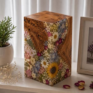 May include: A rectangular wooden urn featuring a natural burl wood grain section integrated with a clear resin panel filled with a variety of pressed dried flowers, including a prominent yellow sunflower, white daisies, and small pink and blue blossoms.