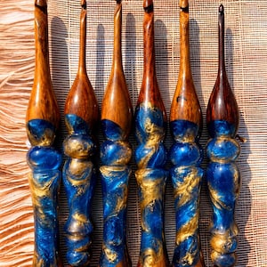 Può includere: Sei uncinetti fatti a mano con manici in legno e dettagli in resina blu e oro. Gli uncinetti sono disposti in fila, mostrando l'esclusiva venatura del legno e i motivi a spirale della resina. Una bella miscela di elementi naturali e artistici.