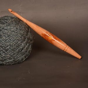 Puede incluir: Un ganchillo de madera con un acabado marrón claro y una madeja de hilo gris. El ganchillo tiene un diseño cónico y un extremo curvado para enganchar el hilo. El hilo es de color gris oscuro con un aspecto texturizado.
