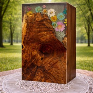 Op de afbeelding: Een houten kist met een natuurlijk houtnerfpatroon. De voorkant heeft een groot, getextureerd houten gedeelte met een knoop. Het bovenste gedeelte is gevuld met heldere hars en kleurrijke gedroogde bloemen. De kist wordt gepresenteerd op een wit kanten kleedje.