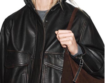 May include: A black leather jacket with a high collar, a front zipper, and two large flap pockets on the chest. The jacket is worn over a grey sweater and is paired with a dark brown suede shoulder bag featuring a silver zipper pull detail.