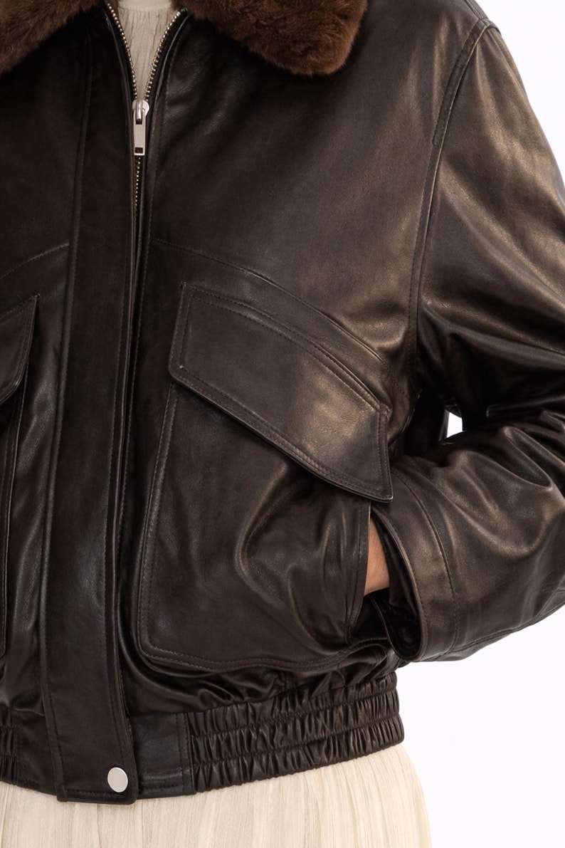 May include: A dark brown leather bomber jacket with a brown fur collar. The jacket features a zip-up front, two front pockets, and a ribbed elastic waistband. The person's hand is in the pocket.
