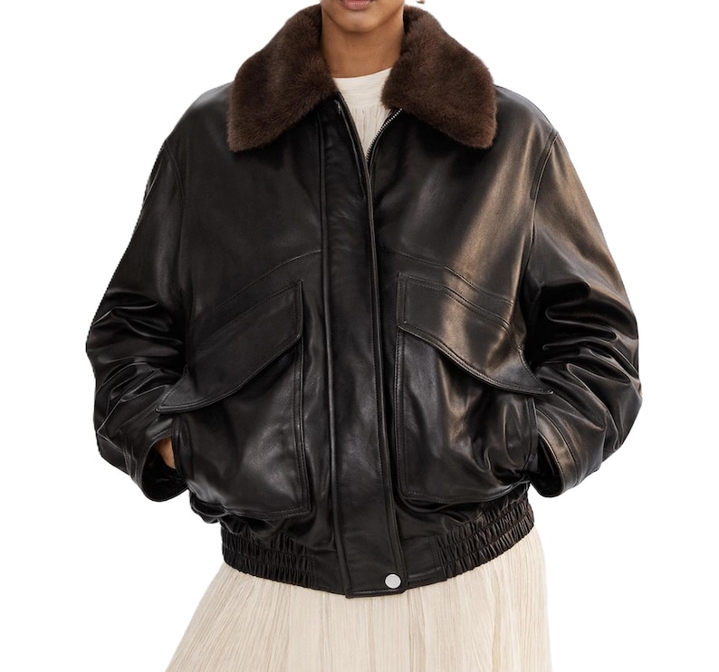May include: A black leather bomber jacket with a brown faux fur collar. The jacket has two front pockets, a zipper closure, and a ribbed hem. The model is wearing a white top and a cream-colored skirt.
