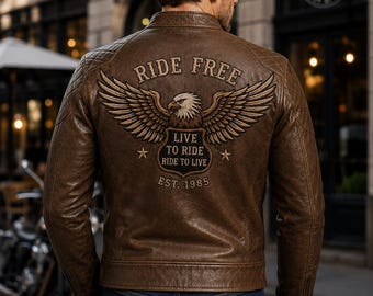Może przedstawiać: Brązowa skórzana kurtka z haftem na plecach przedstawiającym orła oraz napisami RIDE FREE, LIVE TO RIDE, RIDE TO LIVE i EST. 1985. Kurtka posiada pikowane panele na ramionach, stójkę i zamki błyskawiczne przy mankietach, założona na niebieskie dżinsy.