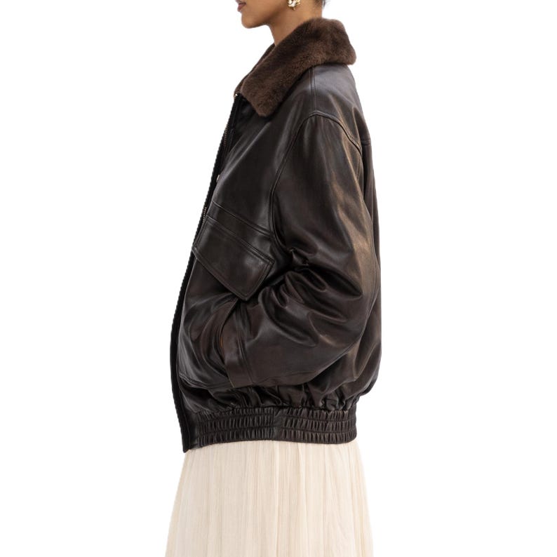 May include: A dark brown leather bomber jacket with a brown fur collar. The jacket has a ribbed hem and cuffs, a zipper closure, and a pocket on the left side. The jacket is a classic style.
