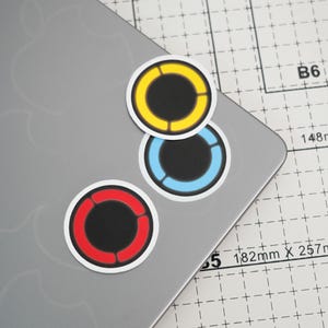 以下が含まれることがあります： 中央が黒く、3つのセグメントに分かれたカラーリングで囲まれた3枚の円形ステッカー。ステッカーは黄色、水色、赤色で、それぞれに細い白い縁取りがあり、グリッド線が引かれた灰色の表面に配置されています。
