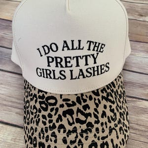 Puede incluir: Gorra de béisbol blanca con el texto "I DO ALL THE PRETTY GIRLS LASHES" bordado en negro. La visera presenta un diseño de estampado de leopardo beige y negro. Un accesorio de moda.