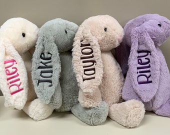 May include: Four plush bunny rabbits in different colors, each with a name embroidered on the ear. The bunnies are light pink, gray, beige, and purple. The names are Riley, Jake, Taylor, and Riley.