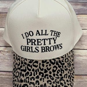 May include: A white baseball cap with the embroidered text "I DO ALL THE PRETTY GIRLS BROWS." The brim features a tan and black leopard print. The cap has a small, decorative button on top.