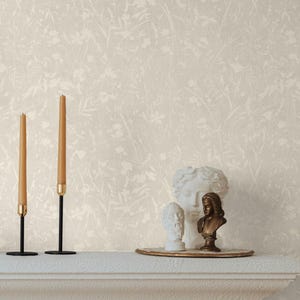 Op de afbeelding: Een crèmekleurige schoorsteenmantel toont twee lange, bruine kaarsen met gouden voetjes en een decoratief dienblad met witte en bronzen bustes. De achtergrond is een crèmekleurig bloemenbehang.