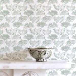 May include: A ceramic bowl with a speckled, textured surface sits on a white shelf. The background features a repeating pattern of light green ginkgo leaves on a white background. The bowl is a neutral color, complementing the soft tones of the wallpaper.
