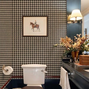 May include: A bathroom interior featuring black and white houndstooth wallpaper, a white toilet with a dark brown seat, a dark vanity with a white towel, a wall-mounted lamp, and a framed print of a person riding a brown horse on the wall.