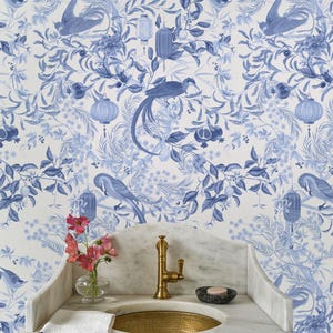 May include: A bathroom sink with a gold faucet and a marble countertop. The wallpaper features a blue and white floral and bird pattern. A small vase of pink flowers and a striped towel add color.