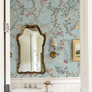 May include: A bathroom with a light blue wallpaper featuring a floral and bird design. A gold-framed mirror hangs above a white marble sink. A small framed picture and a sconce with a beige lampshade are also visible.
