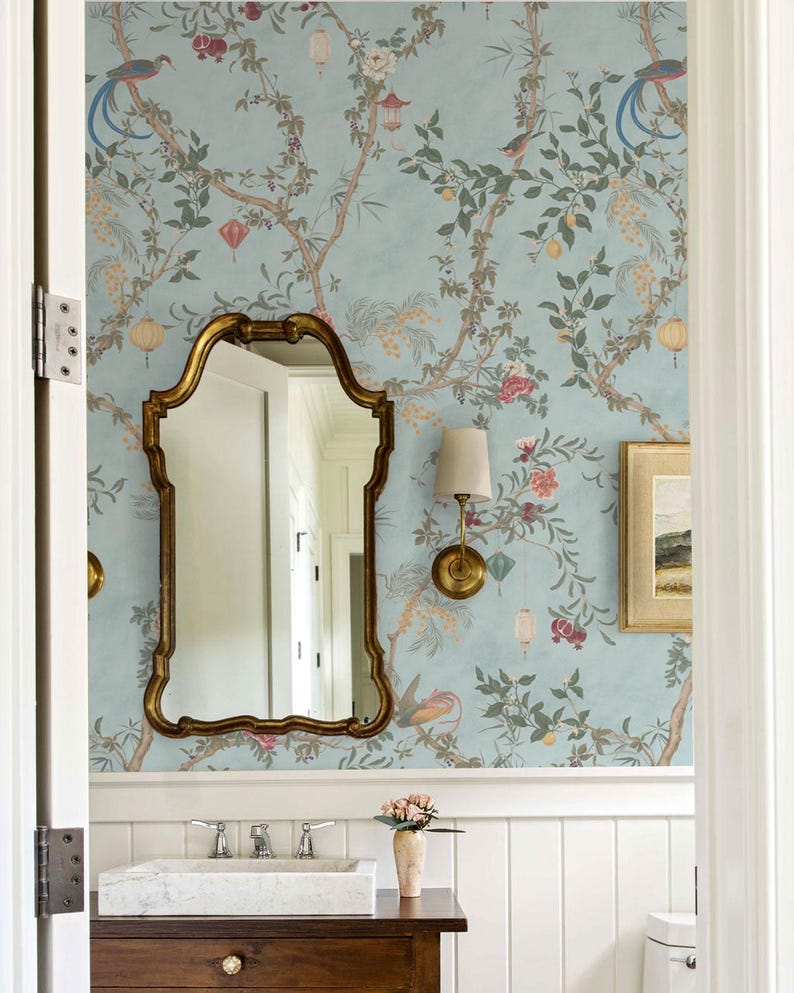 May include: A bathroom with a light blue wallpaper featuring a floral and bird design. A gold-framed mirror hangs above a white marble sink. A small framed picture and a sconce with a beige lampshade are also visible.