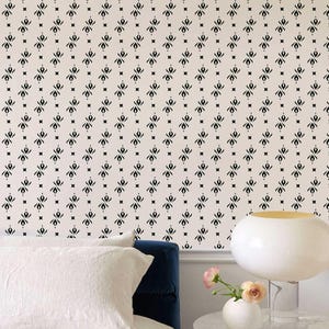 May include: A bedroom scene featuring a cream-colored wall with a repeating black geometric pattern. A white pillow and a blue headboard are visible. A white lamp with a gold interior and a small vase with a pink flower sit on a marble table.