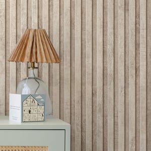 May include: A table lamp with a tan, pleated lampshade sits on a light blue cabinet. A clear glass base supports the lamp. A greeting card with a house illustration is on the cabinet. The background is a vertical wood-paneled wall.