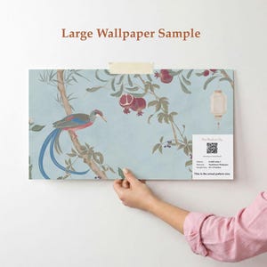 May include: A large wallpaper sample featuring a bird perched on a branch with pomegranates and a lantern against a light blue background. The sample is held up against a white wall. Text on the sample reads "Large Wallpaper Sample" and "This is the actual pattern size."