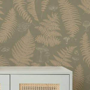May include: A close-up of a wallpaper design featuring a repeating pattern of ferns and mushrooms in shades of tan and white on a muted olive green background. The bottom of a light blue cabinet with a woven rattan panel is visible.