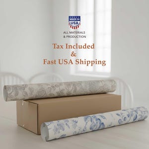 May include: Two rolls of floral wallpaper rest on a cardboard box. One roll features a gray floral pattern, while the other displays a blue floral design. The image also includes text that reads "Made in USA" and "All Materials & Production."