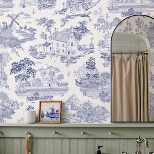 May include: Blue and white toile wallpaper featuring a pastoral landscape with buildings, trees, and water elements. A framed picture of a windmill scene and a white ceramic cup sit on a green shelf. A mirror and shower curtain are also visible.