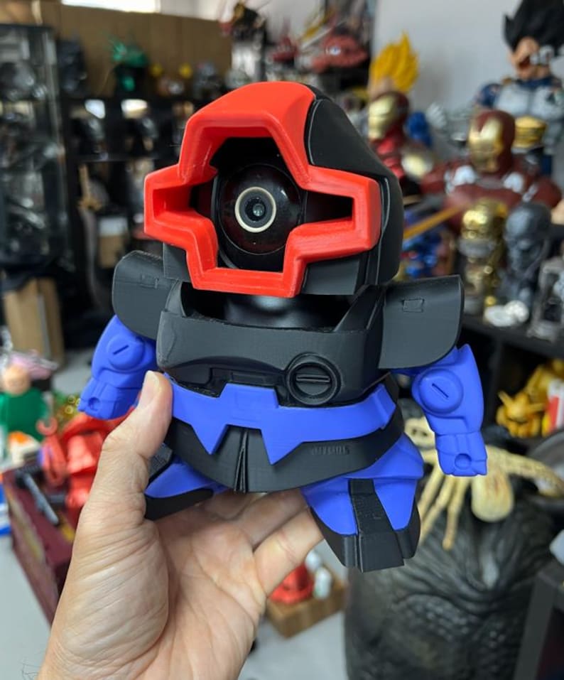 Puede incluir: Un juguete robot impreso en 3D con una combinaci&oacute;n de colores rojo, negro y azul. El robot tiene un ojo de c&aacute;mara y una visera roja en forma de cruz. El juguete se sostiene en una mano, con otras figuras visibles en el fondo.