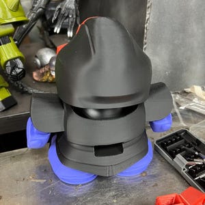 Puede incluir: Una figura impresa en 3D con casco y cuerpo negros, piernas azules y detalles rojos. La figura est&aacute; sobre una superficie met&aacute;lica. Otros objetos son visibles en el fondo, incluyendo una figura verde y una figura negra.