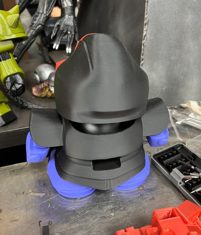 Puede incluir: Una figura impresa en 3D con casco y cuerpo negros, piernas azules y detalles rojos. La figura est&aacute; sobre una superficie met&aacute;lica. Otros objetos son visibles en el fondo, incluyendo una figura verde y una figura negra.