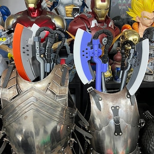 Op de afbeelding: Twee harnassen met bijlen. E&eacute;n bijl heeft een oranje blad, de andere een wit blad. Het harnas is zilverkleurig en heeft leren riemen. Standbeelden van Iron Man en andere personages staan op de achtergrond.