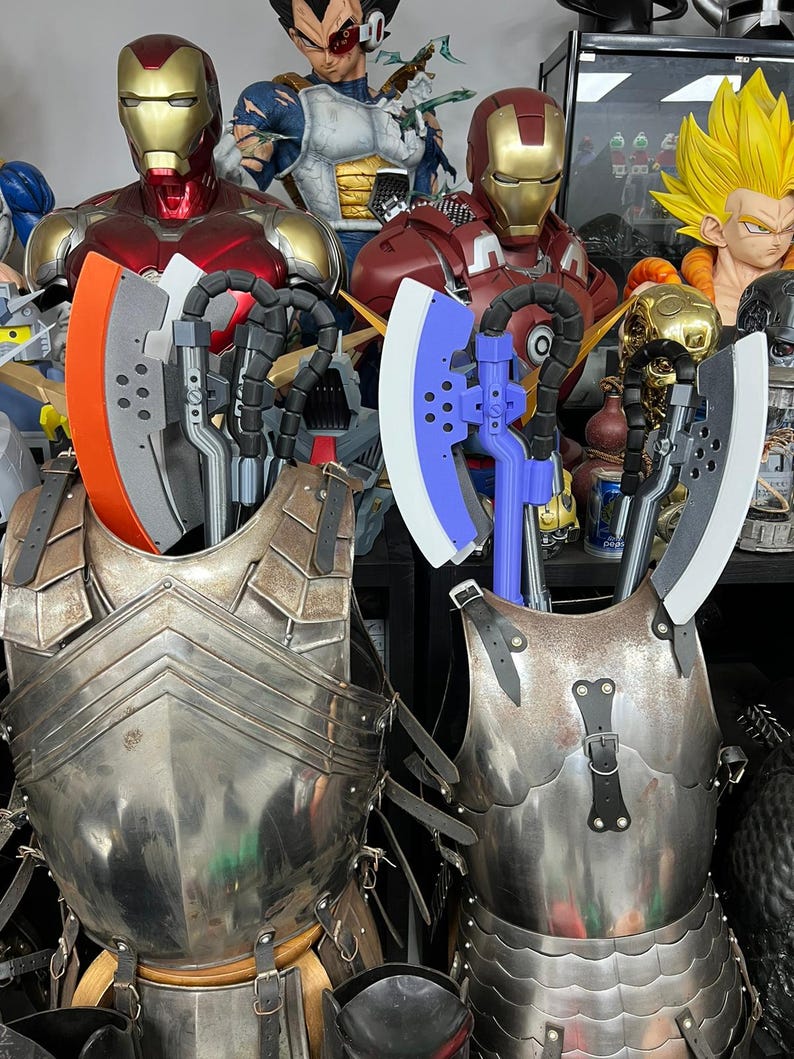 Op de afbeelding: Twee harnassen met bijlen. E&eacute;n bijl heeft een oranje blad, de andere een wit blad. Het harnas is zilverkleurig en heeft leren riemen. Standbeelden van Iron Man en andere personages staan op de achtergrond.