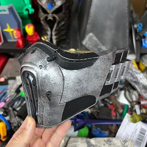 May include: A silver and black mask with a distressed finish. The mask features intricate detailing, including layered elements and geometric patterns. The mask is held in a hand, indicating it's a wearable item, likely for cosplay or display purposes.