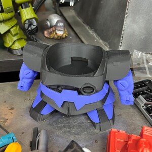Puede incluir: Un modelo de robot impreso en 3D en negro y azul, con brazos y piernas azules. El robot tiene una abertura circular en la parte superior y est&aacute; colocado sobre una superficie met&aacute;lica. Otras piezas de juguete son visibles en el fondo.