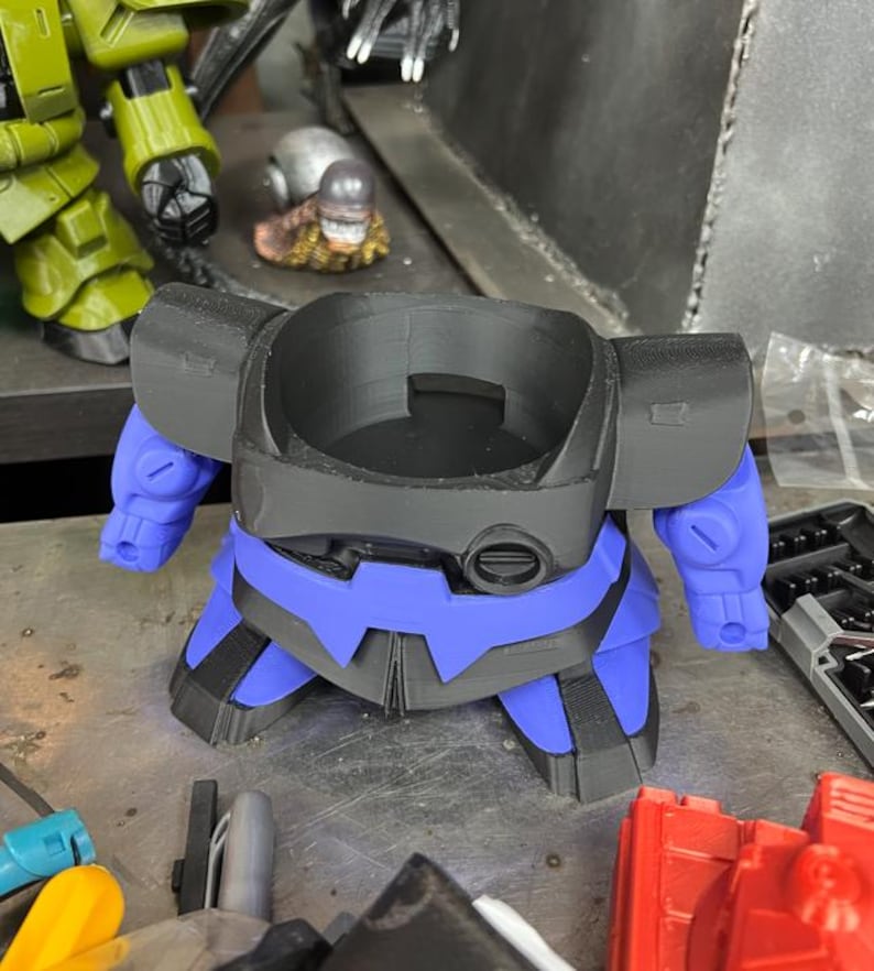 Puede incluir: Un modelo de robot impreso en 3D en negro y azul, con brazos y piernas azules. El robot tiene una abertura circular en la parte superior y est&aacute; colocado sobre una superficie met&aacute;lica. Otras piezas de juguete son visibles en el fondo.