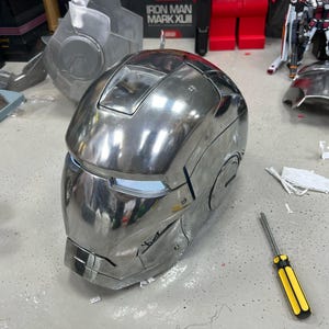 Puede incluir: Un casco de Iron Man metálico pulido y brillante. El casco es plateado con una abertura rectangular para los ojos y un diseño detallado. Un destornillador amarillo y negro está cerca. Otras piezas desmontadas están en el fondo.