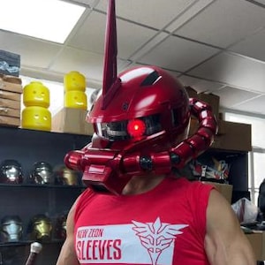 May include: A metallic red Zaku-style helmet featuring a prominent vertical antenna, a singular glowing red circular eye, and a segmented tube extending from the side. The helmet is worn above a red sleeveless shirt with the text NEW ZEON SLEEVES.