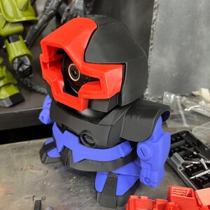 Puede incluir: Un juguete robot impreso en 3D con cabeza y torso negros, visera roja y brazos y piernas azules. El robot tiene una lente de c&aacute;mara circular en el centro de su cara. Otras piezas de juguete son visibles en el fondo.