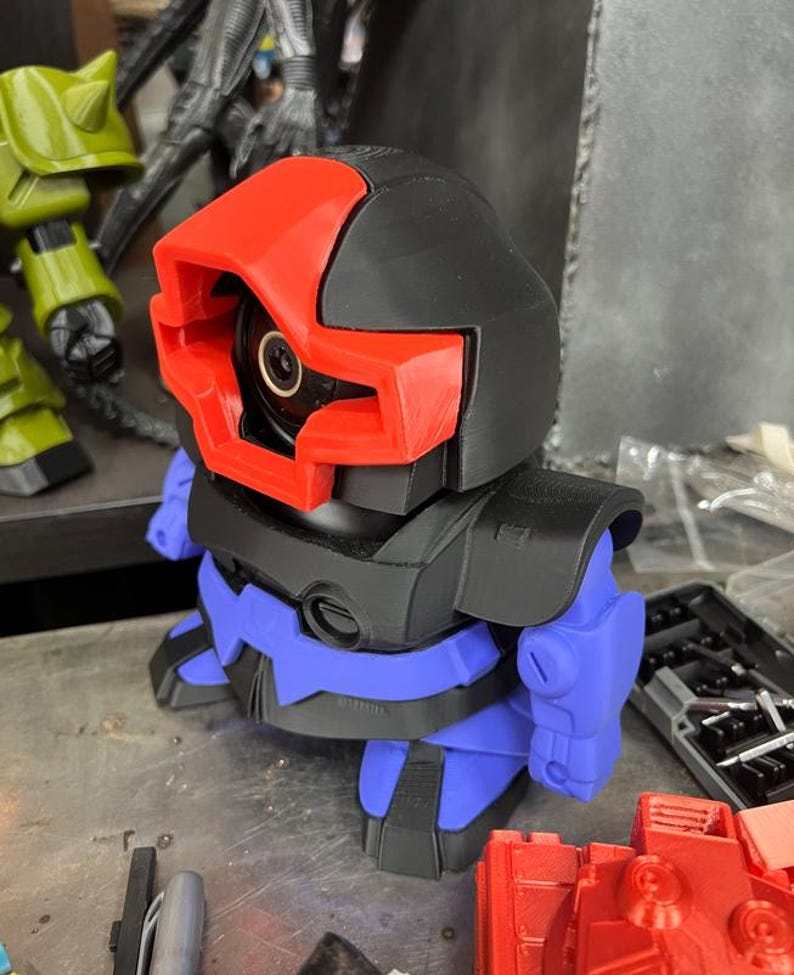 Puede incluir: Un juguete robot impreso en 3D con cabeza y torso negros, visera roja y brazos y piernas azules. El robot tiene una lente de c&aacute;mara circular en el centro de su cara. Otras piezas de juguete son visibles en el fondo.