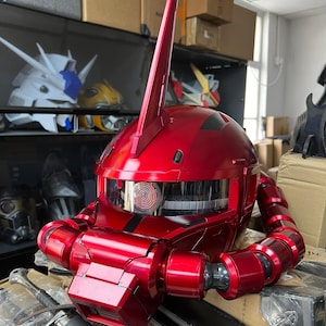 以下が含まれることがあります： 光沢のある赤いロボットヘルメット。中央に目立つフィンがあり、詳細な機械的特徴を備えています。ヘルメットは透明なバイザーと関節式の首部分を備えています。他のロボットヘルメットが背景に見えます。