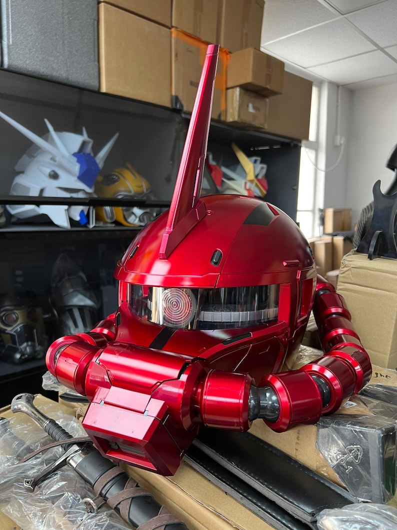 以下が含まれることがあります： 光沢のある赤いロボットヘルメット。中央に目立つフィンがあり、詳細な機械的特徴を備えています。ヘルメットは透明なバイザーと関節式の首部分を備えています。他のロボットヘルメットが背景に見えます。