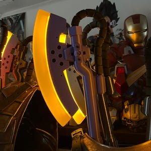 Op de afbeelding: Een close-up van een fantasiebijl met een gloeiend geel blad en een paarse kop. De bijl heeft een gedetailleerd ontwerp met een metalen handvat. Onderdeel van een verzameling fantasie-items.